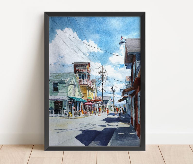 May include: Watercolor painting of a coastal street scene with buildings in shades of green, yellow, and red. A blue sky with fluffy clouds is above. People are walking on the street. The artwork is framed in black.