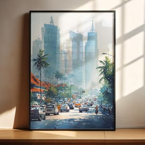 May include: A framed watercolor print of a city street scene. Tall, light blue skyscrapers rise in the background, with palm trees and traffic in the foreground. The artwork is framed in a black border and is displayed on a wooden surface.