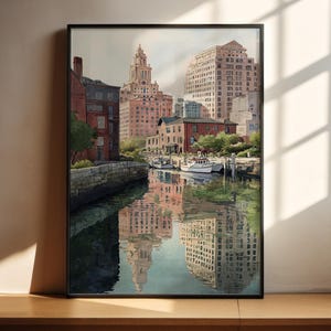 May include: A framed watercolor painting of a city harbor scene. The artwork depicts buildings in shades of red, beige, and brown, reflected in the water. A few boats are docked near the buildings. The sky is overcast, and the painting is in a black frame.