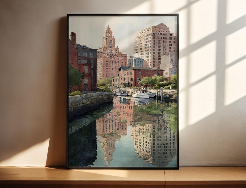 May include: A framed watercolor painting of a city harbor scene. The artwork depicts buildings in shades of red, beige, and brown, reflected in the water. A few boats are docked near the buildings. The sky is overcast, and the painting is in a black frame.