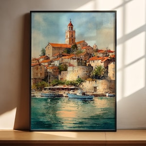 May include: Watercolor painting of a coastal town with red-tiled roofs, a tall bell tower, and boats on the water. The artwork is framed in a black border and set against a neutral wall. The water reflects the sunlight, creating a serene atmosphere.