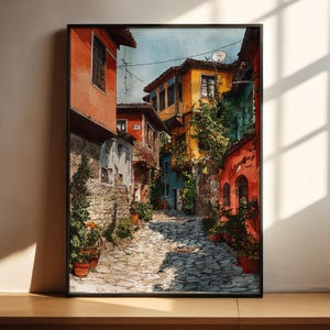 May include: Watercolor painting of a European street scene with colorful buildings in shades of red, orange, yellow, and blue. The cobblestone street is lined with potted plants. The artwork is framed in a black frame.