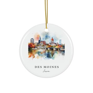 May include: A round white ceramic ornament featuring a colorful watercolor illustration of the Des Moines, Iowa skyline with a bridge over water. The text DES MOINES Iowa is printed in black below the city scene. A gold string is attached at the top.