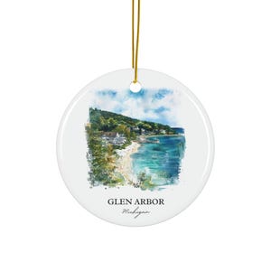 Könnte beinhalten: Runder weißer Keramikschmuck mit einem Aquarellgemälde von Glen Arbor, Michigan. Das Bild zeigt einen Strand, Wasser und Gebäude unter blauem Himmel. Der Text "Glen Arbor Michigan" ist auf dem Ornament aufgedruckt.