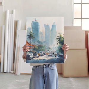 May include: A watercolor painting of a city street scene with tall buildings, palm trees, and traffic. The artwork is held by a person in a studio setting. The painting features a color palette of blues, greens, and oranges.