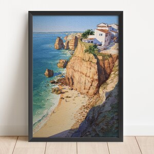 May include: Framed watercolor painting of a coastal landscape. The artwork depicts a beach with cliffs, ocean, and buildings. The cliffs are a mix of tan and brown, and the ocean is blue. The buildings are white with red roofs.