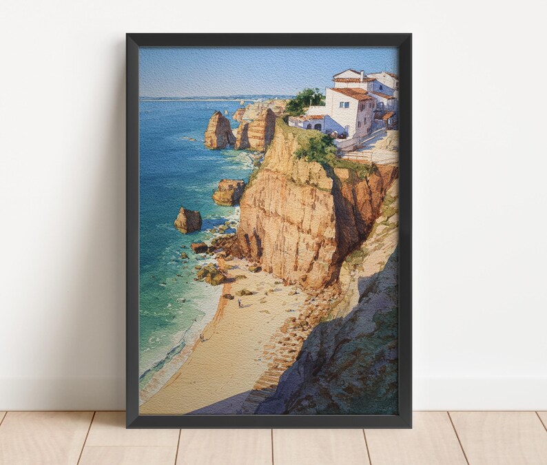 May include: Framed watercolor painting of a coastal landscape. The artwork depicts a beach with cliffs, ocean, and buildings. The cliffs are a mix of tan and brown, and the ocean is blue. The buildings are white with red roofs.
