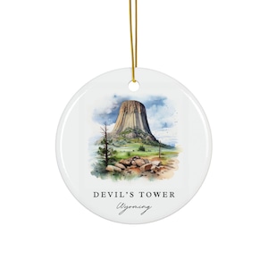 May include: White ceramic ornament featuring a watercolor illustration of Devil's Tower in Wyoming. The image shows the tower against a cloudy sky, with text reading "DEVIL'S TOWER Wyoming". A gold string is attached for hanging.
