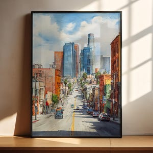 May include: Watercolor painting of a city street scene with tall buildings and palm trees. The artwork features a black frame and captures a sunny day with a blue sky and clouds. Cars are visible on the road, and the buildings are in shades of orange, blue, and brown.