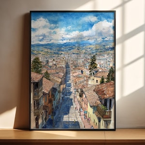 May include: Watercolor painting of a city street scene, framed in black. The artwork depicts a long street lined with buildings, leading towards a distant mountain range under a blue sky with fluffy clouds. The buildings have red tile roofs.