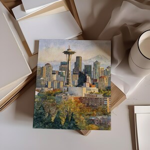 May include: Watercolor painting of the Seattle skyline, featuring the Space Needle and surrounding buildings. The artwork is displayed on a white surface, with additional white paper and a decorative cup in the background. The painting captures the city's architecture and natural surroundings.