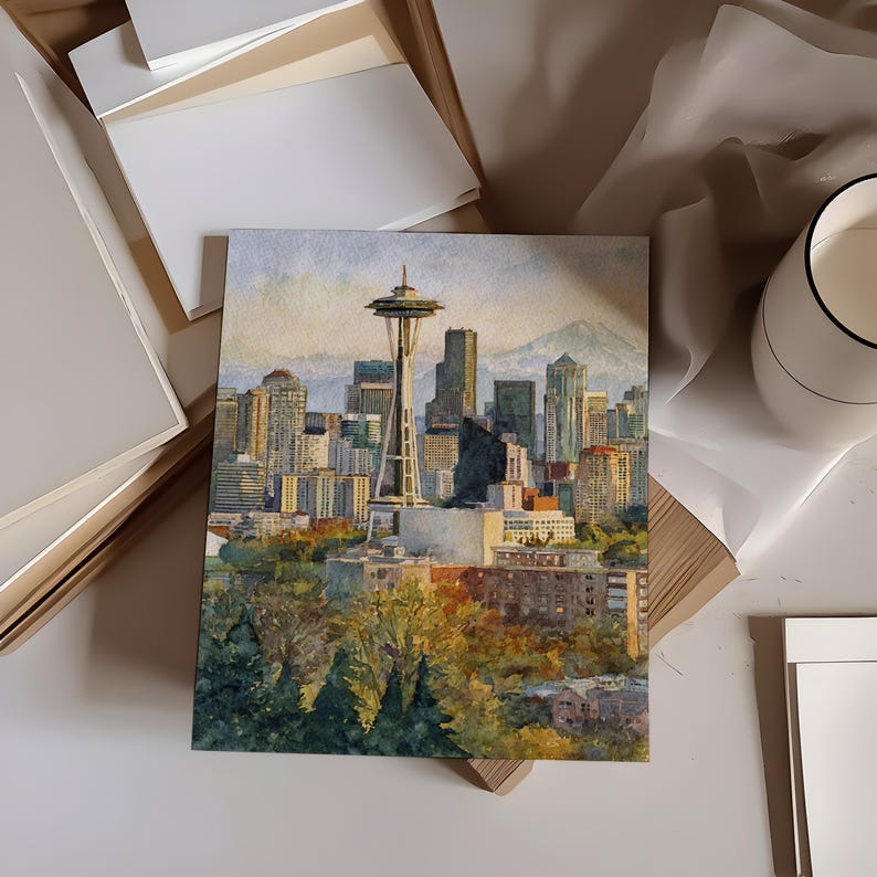 May include: Watercolor painting of the Seattle skyline, featuring the Space Needle and surrounding buildings. The artwork is displayed on a white surface, with additional white paper and a decorative cup in the background. The painting captures the city's architecture and natural surroundings.
