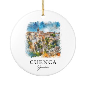 May include: A round white ceramic ornament featuring a watercolor illustration of the city of Cuenca, Spain, perched on cliffs. The text CUENCA Spain is printed in black below the image. A gold string is attached to the top for hanging.