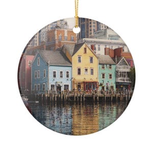 Può includere: Un ornamento rotondo in ceramica con un'illustrazione dettagliata di edifici colorati sul lungomare in blu, giallo e verde acqua, posizionati su pali di legno sopra l'acqua, con un cordino dorato in cima per appenderlo a un supporto o albero.
