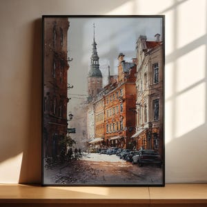 May include: Watercolor painting of a European city street scene. The artwork depicts buildings with red-brown facades, a tall church spire, and parked cars. The painting is framed in a black border and rests on a wooden surface.