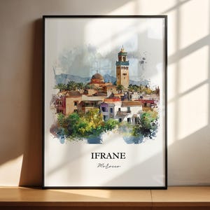 Könnte beinhalten: Ein Aquarellbild einer Stadtlandschaft mit einem hohen Minarett und anderen Gebäuden. Das Bild ist in Schwarz gerahmt und trägt den Schriftzug "IFRANE" und "Marokko" unterhalb des Bildes.
