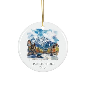 May include: White ceramic ornament with a watercolor painting of Jackson Hole, Wyoming. The design features a street scene with buildings, cars, and snow-capped mountains under a blue sky. The text "JACKSON HOLE Wyoming" is printed below the image.