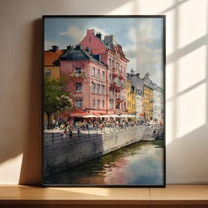 Puede incluir: Pintura a la acuarela de una escena de ciudad europea con edificios coloridos que bordean un canal. Los edificios son rosas, amarillos y grises, con toldos rojos sobre zonas de estar al aire libre. Se ve un árbol y personas.