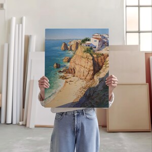 May include: A landscape painting depicts a coastal scene with cliffs, a sandy beach, and buildings. The artwork features a vibrant blue ocean, golden cliffs, and white buildings under a clear sky. The painting is held by a person.
