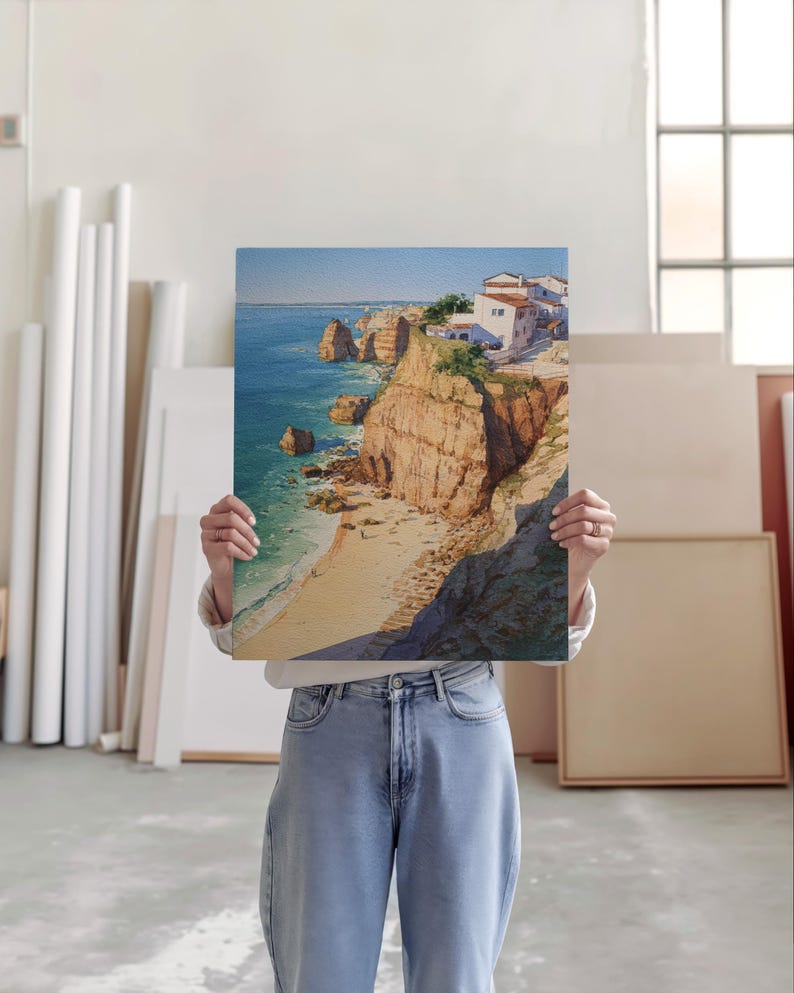May include: A landscape painting depicts a coastal scene with cliffs, a sandy beach, and buildings. The artwork features a vibrant blue ocean, golden cliffs, and white buildings under a clear sky. The painting is held by a person.