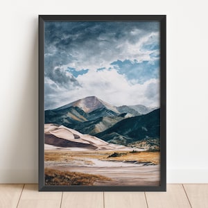 May include: Framed watercolour landscape art print. The artwork depicts a mountain range with sand dunes in the foreground. The sky is filled with dramatic clouds. The colour palette includes blues, greens, browns, and beige.