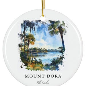 May include: White ceramic ornament with a watercolor painting of a lake scene with palm trees and the text "MOUNT DORA Florida". The ornament has a gold string for hanging.