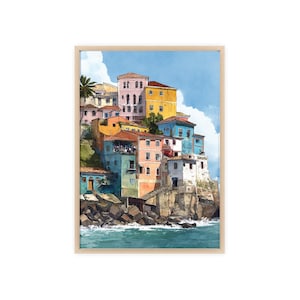 May include: Watercolor painting of a coastal village with colorful buildings. The buildings are stacked on a rocky cliffside, overlooking the ocean. The artwork is framed in a light wood frame. The sky is blue with fluffy white clouds.