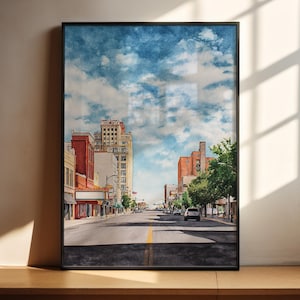 May include: A framed watercolor painting of a city street scene. The artwork depicts a wide road with buildings on either side, under a blue sky with fluffy white clouds. The buildings are a mix of red brick and beige tones.
