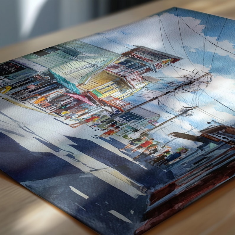 May include: Watercolor painting of a street scene with buildings and people. The artwork features a vibrant color palette with blues, yellows, and reds. The composition includes a street with shadows, power lines, and a bright blue sky with clouds.