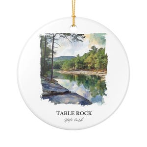 Puede incluir: Adorno redondo de cerámica blanca con una pintura de acuarela del Parque Estatal Table Rock. La imagen presenta un lago, árboles y montañas. El texto "TABLE ROCK State Park" está impreso en negro en la parte inferior, con una cuerda dorada para colgar.