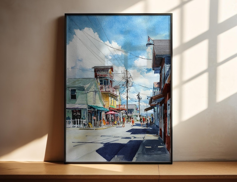 May include: Watercolor painting of a street scene with buildings and people. The artwork features a blue sky with clouds, a street with shadows, and colorful buildings. The painting is framed in a black border.