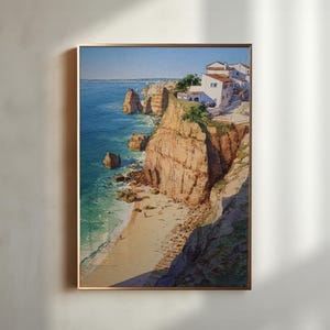 May include: A framed watercolor painting depicts a coastal scene with cliffs, a beach, and buildings. The artwork features a vibrant blue ocean, tan cliffs, and white buildings with red tile roofs. The painting is framed in a gold-colored frame.