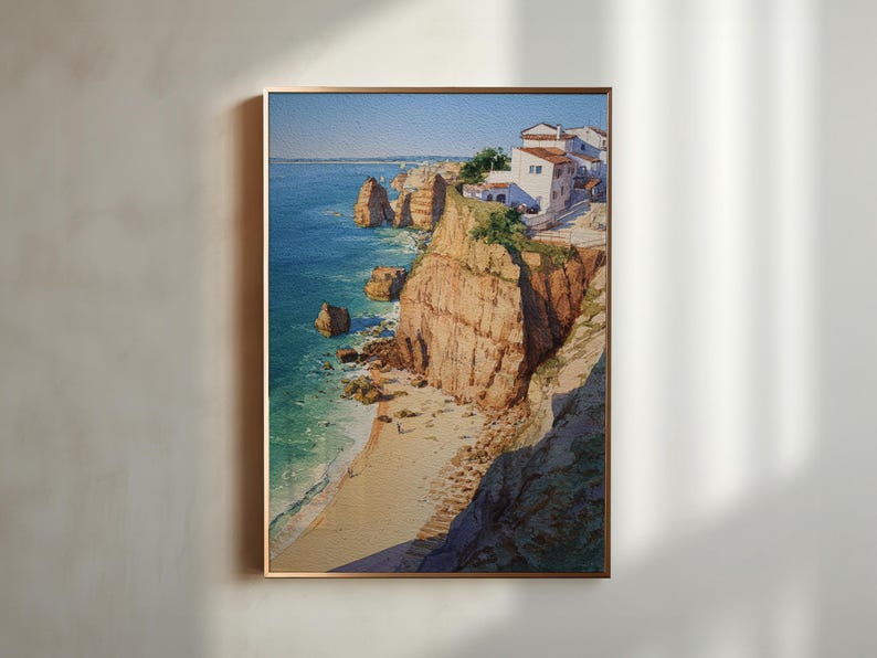 May include: A framed watercolor painting depicts a coastal scene with cliffs, a beach, and buildings. The artwork features a vibrant blue ocean, tan cliffs, and white buildings with red tile roofs. The painting is framed in a gold-colored frame.