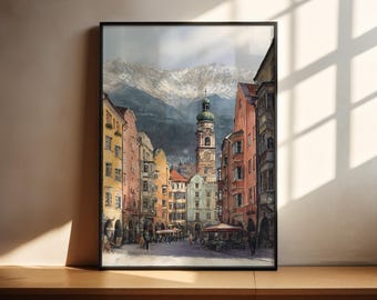 Könnte beinhalten: Ein gerahmter Aquarelldruck einer europäischen Stadtstraßenszene. Das Kunstwerk zeigt farbenfrohe Gebäude, einen Glockenturm und schneebedeckte Berge im Hintergrund. Der Druck wird auf einer Holzoberfläche präsentiert.