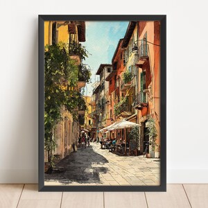 May include: Framed watercolor print of a European street scene. The artwork depicts a narrow street lined with colorful buildings, balconies, and awnings. People are seated at outdoor tables. The sky is a light blue.