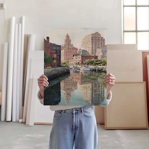 May include: A rectangular art print depicting a cityscape with buildings reflected in the water. The buildings are primarily brick and beige, with a few boats docked along the water's edge. The print is held up in front of a person.
