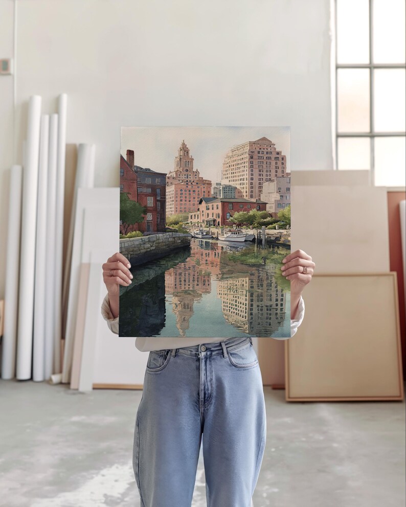 May include: A rectangular art print depicting a cityscape with buildings reflected in the water. The buildings are primarily brick and beige, with a few boats docked along the water's edge. The print is held up in front of a person.