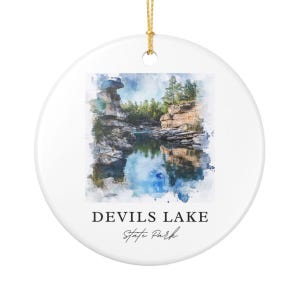 May include: A round white ceramic ornament featuring a watercolor-style illustration of a rocky landscape with a lake and trees. The text "DEVILS LAKE" is printed in black capital letters above the cursive script text "State Park".