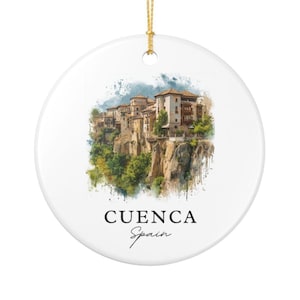 May include: A round white ceramic ornament featuring a watercolor-style illustration of the hanging houses of Cuenca, Spain, perched on a cliffside. The text CUENCA Spain is printed in black below the image, and a gold string is attached at the top.