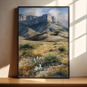 May include: A framed landscape print featuring a mountain range under a cloudy sky. The artwork depicts rolling hills and desert vegetation in shades of green, brown, and yellow. The print is displayed in a black frame and rests on a wooden surface.