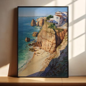 May include: A framed watercolor print of a coastal scene. The artwork depicts a cliffside with white buildings, a sandy beach, and turquoise water. The cliffs are a mix of tan and brown, and the sky is a clear blue.