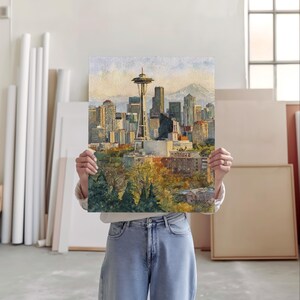 May include: Watercolor painting of the Seattle skyline, including the Space Needle, skyscrapers, and a mountain in the background. The artwork features warm colors and a detailed depiction of the city's architecture and natural surroundings. The painting is held by a person.