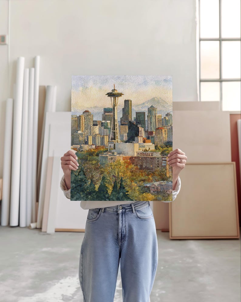 May include: Watercolor painting of the Seattle skyline, including the Space Needle, skyscrapers, and a mountain in the background. The artwork features warm colors and a detailed depiction of the city's architecture and natural surroundings. The painting is held by a person.