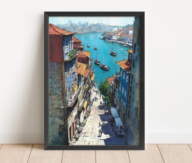 Puede incluir: Una pintura de acuarela enmarcada representa un paisaje urbano europeo con edificios que bordean una calle que conduce a un r&iacute;o. Los edificios tienen tejados de tejas rojas y fachadas coloridas. Hay barcos en el agua azul. La obra de arte est&aacute; en un marco negro.