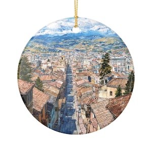 May include: A round ceramic ornament featuring a detailed watercolor painting of a city street lined with buildings that have terracotta-colored roofs, leading toward distant blue mountains under a cloudy sky, suspended by a gold string.