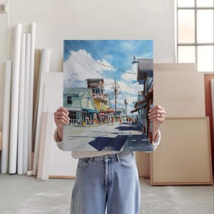 May include: A watercolor painting of a street scene with shops and people. The artwork depicts a sunny day with blue skies and clouds. The buildings are painted in various colors, and the street is lined with people.