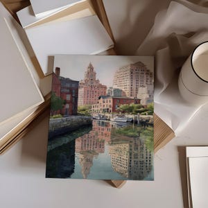 May include: A square watercolor painting depicts a city harbor scene with buildings reflected in the water. The artwork features a mix of red brick and light-colored buildings, with boats docked along the waterfront. The painting is surrounded by art supplies.