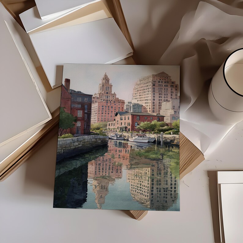 May include: A square watercolor painting depicts a city harbor scene with buildings reflected in the water. The artwork features a mix of red brick and light-colored buildings, with boats docked along the waterfront. The painting is surrounded by art supplies.