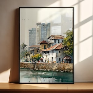 May include: Watercolour painting of a coastal town with buildings, palm trees, and a body of water. The artwork is framed in a black border and displayed on a wooden surface. The painting features a muted colour palette with shades of blue, brown, and green.