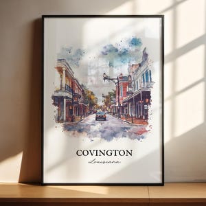 May include: Framed watercolor print of Covington, Louisiana, featuring a street scene with buildings and a car. The artwork has a soft, colorful aesthetic with the city name and state written below.
